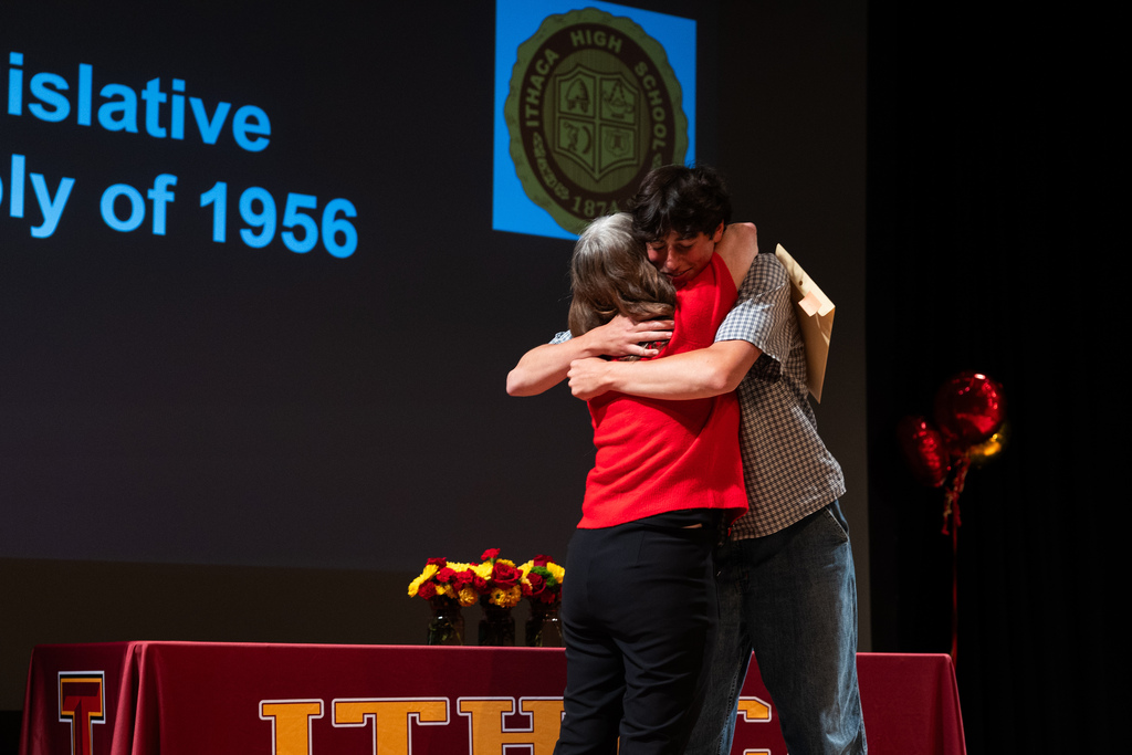 Two people embrace on a stage in front of a screen. A table with a red tablecloth is in front of them.