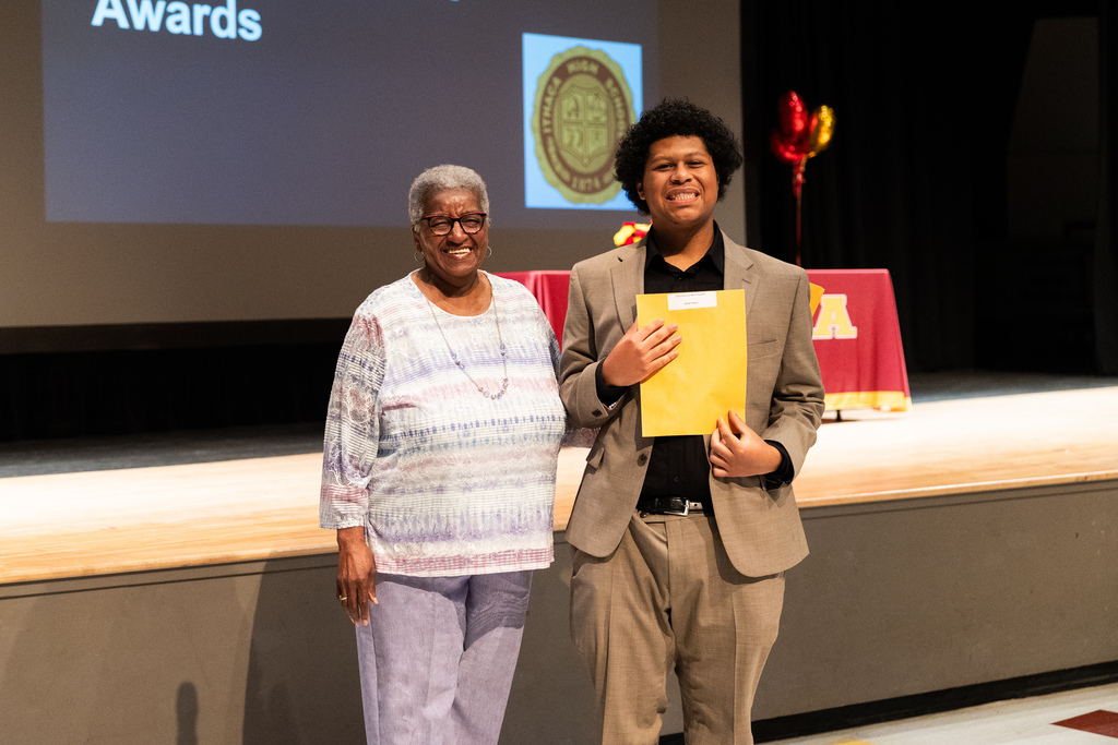 Two individuals, one holding a yellow folder, stand on a stage with a screen behind them displaying the word Awards.