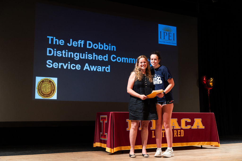 Two women stand on stage with an award; a large screen behind them displays "The Jeff Dobbin Distinguished Community Service Award".