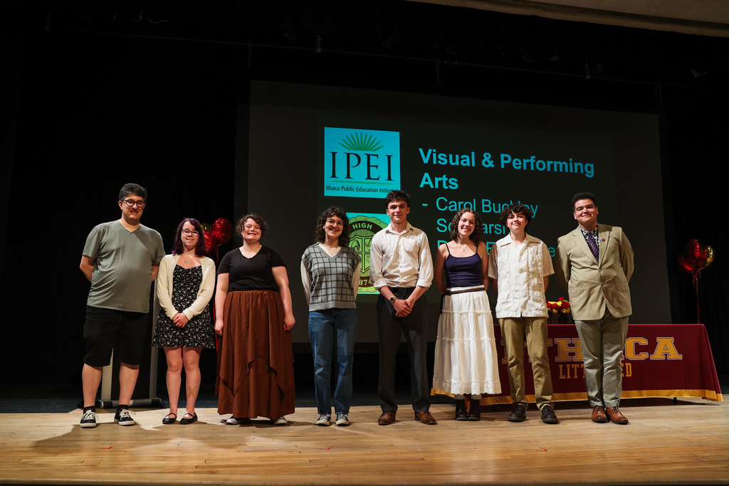 Seven people on a stage with a large screen behind them displaying "Visual & Performing Arts." A table holds red flowers.