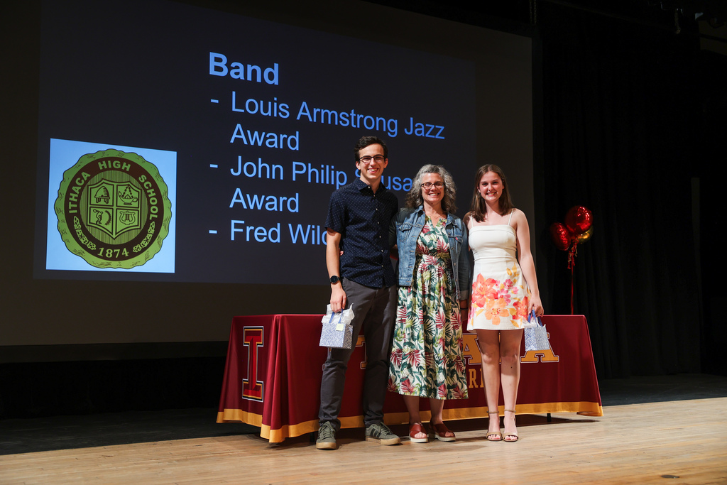 Three people stand on stage holding awards. A presentation screen is behind them. A table with a red cover is in front of them.