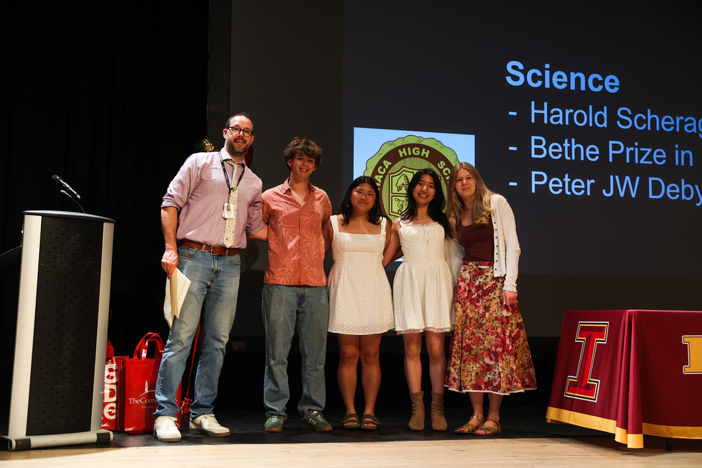 Five people on a stage, one with a microphone. Behind them is a screen with text. An I sign and table nearby.