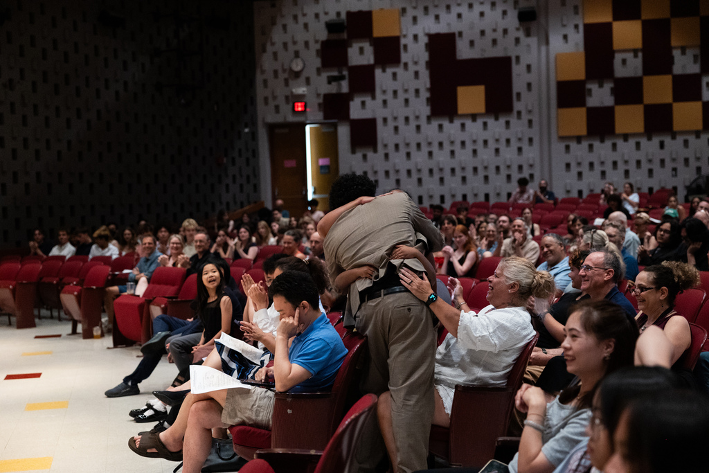 An auditorium with red seats, where several people sit while a person stands in the middle, possibly during an event.