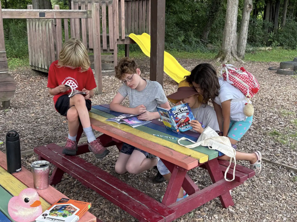 Four children are seated around a picnic table. Two are reading books, and one is holding a book. A slide and trees are in the background.