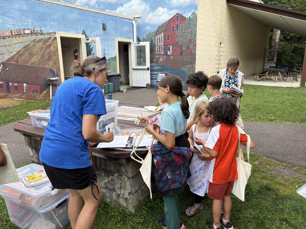 A group of children and a woman examine items on a table in front of a mural.