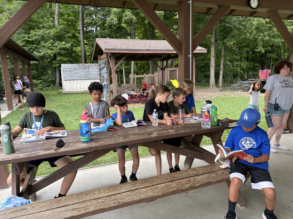 Children at a picnic table, reading, with a woman nearby, a clock on a gazebo in the background.