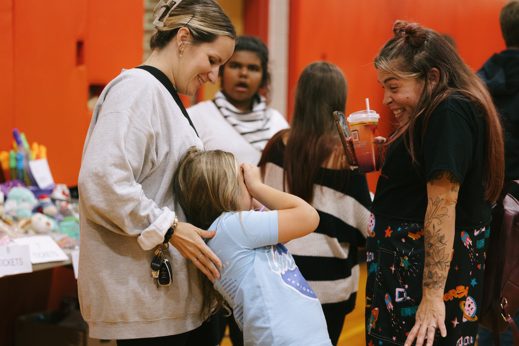 A woman standing behind a student, both looking at a smiling woman; behind them, others talk or hold cups.