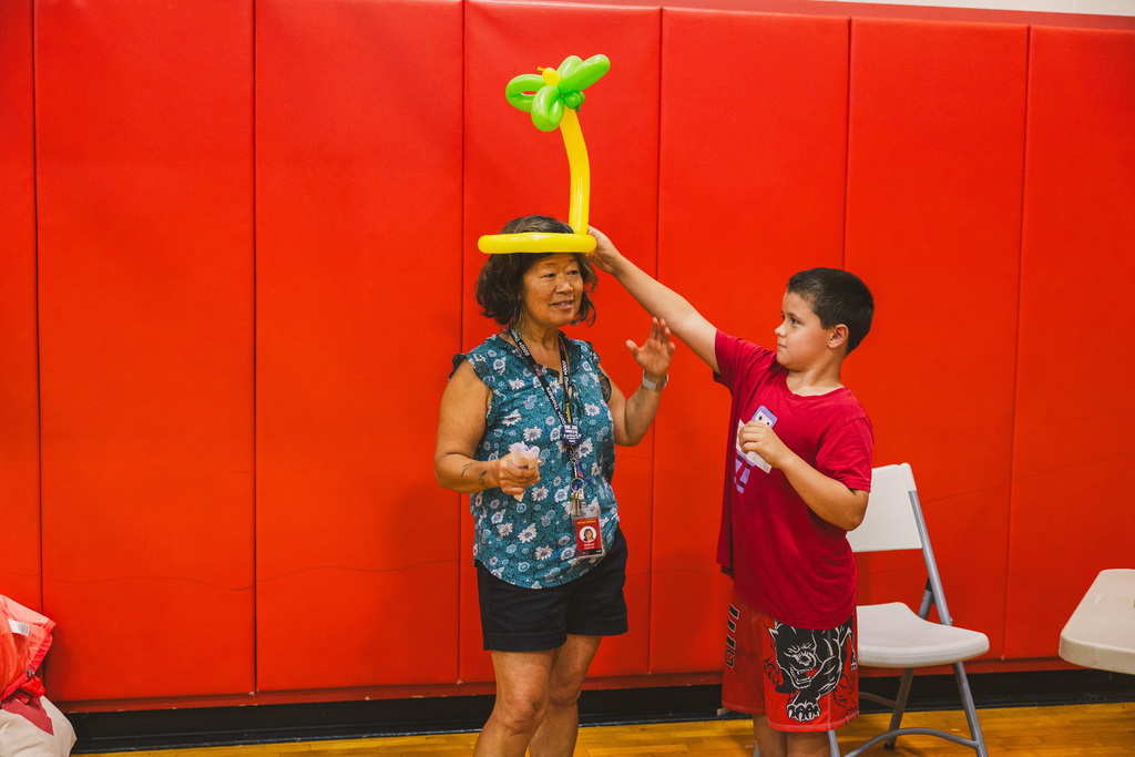 A woman and a child stand next to each other while the child puts a balloon on the woman's head, standing against a red wall.