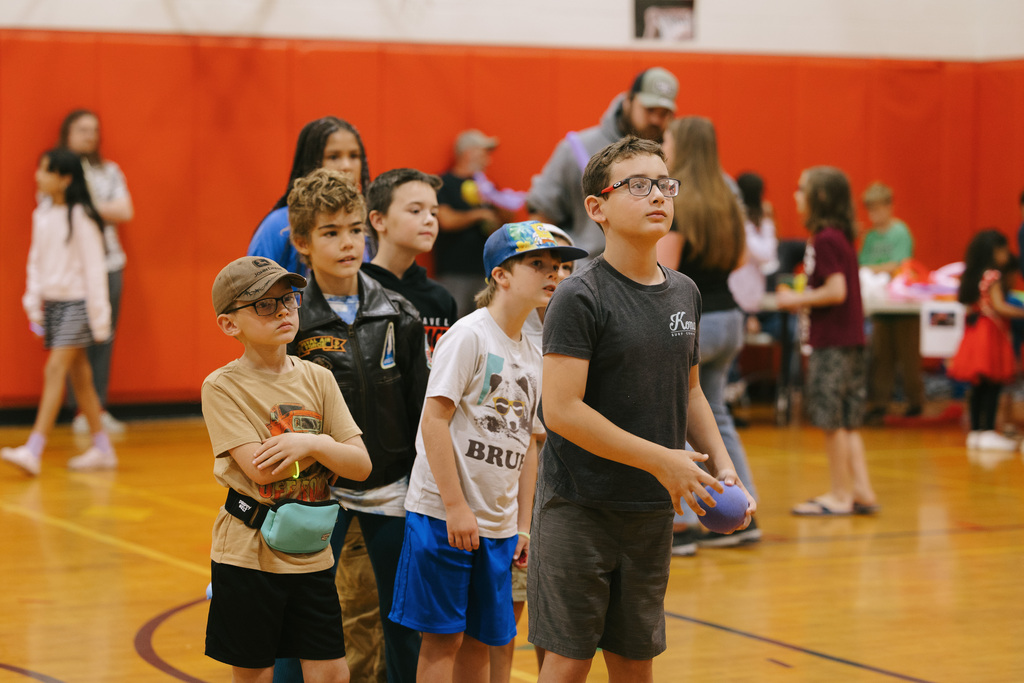 Multiple children and adults are standing in a gymnasium. They are wearing casual clothes and sneakers.