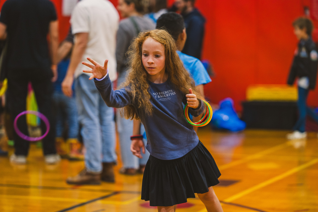 A girl waves rings while standing in a gymnasium. People stand behind her, and a red wall is visible.