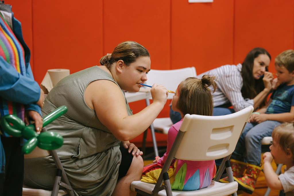 A woman face paints a student in the Enfield gym while other children sit nearby waiting to have their faces painted.