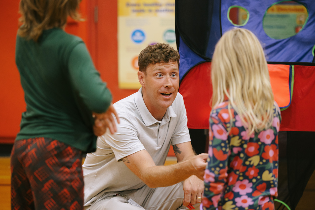 Principal Stephan crouching speaks to a girl. Another child stands nearby. Background contains a multicolored tent.
