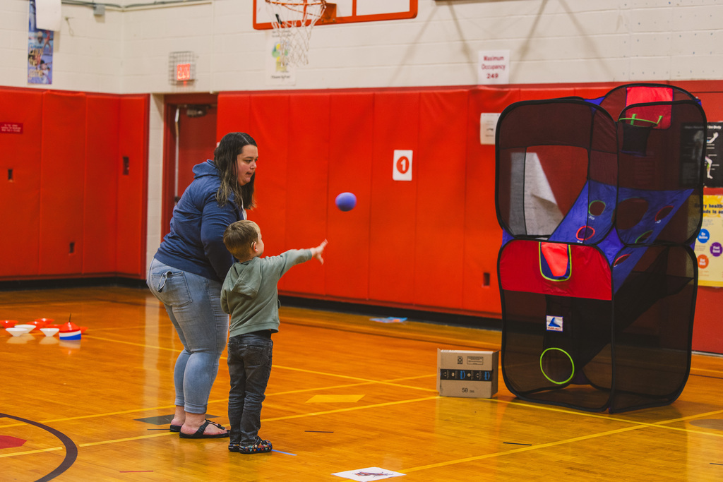 A woman and a young child in a gymnasium. The child throws a blue ball at a pop-up toy.