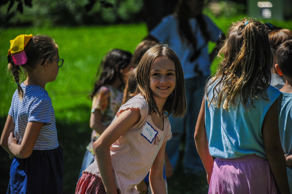 A group of children stand in a grassy area, one smiling. The background features a tree and a person in a white shirt.