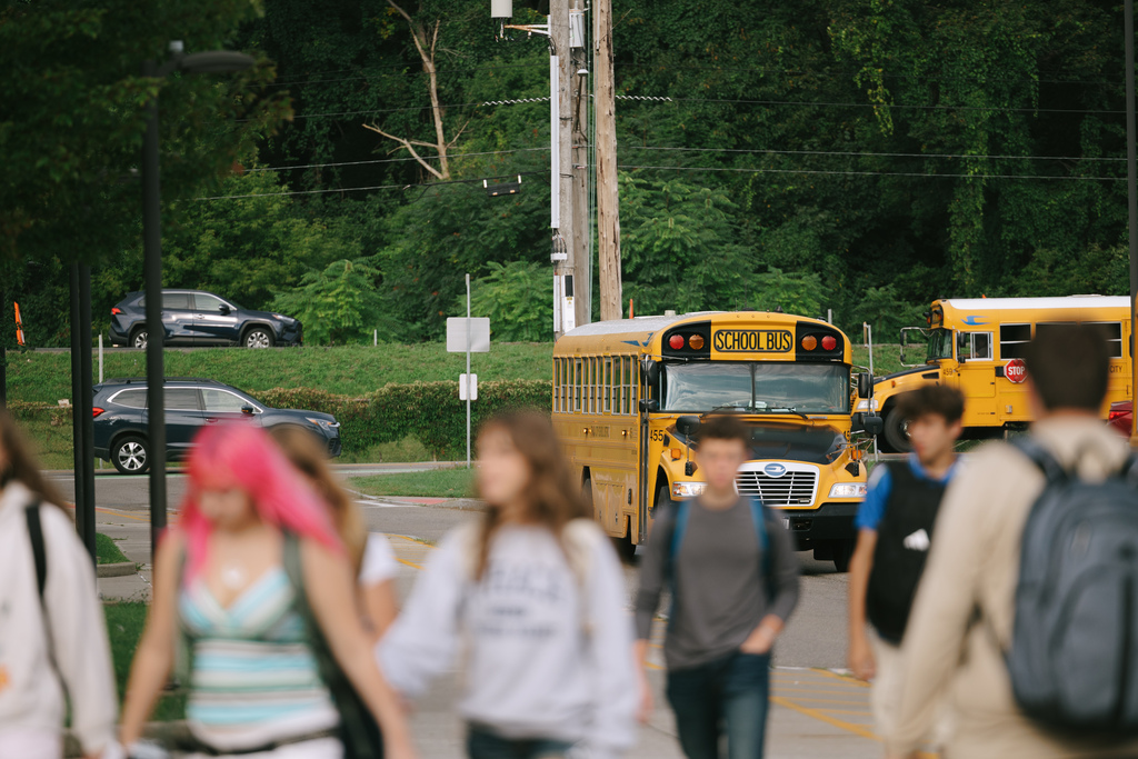 School buses in a parking lot. Some students walk past, some with backpacks. Greenery and trees in background.