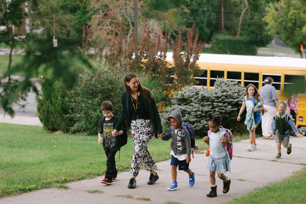 A woman and five children walk on a sidewalk. Greenery and a yellow school bus are in the background.