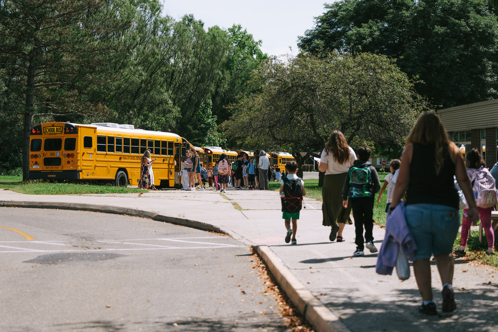 Several children walking down the sidewalk, with yellow school buses parked on the side.