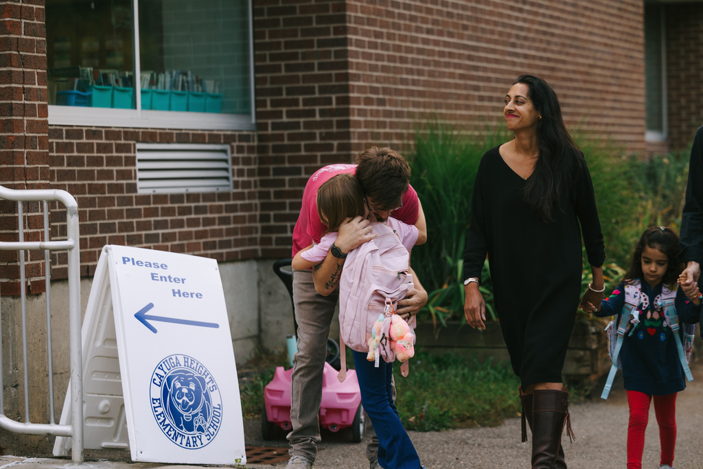 A family walks on a sidewalk with a woman hugging a girl. A sign with an arrow is beside them.