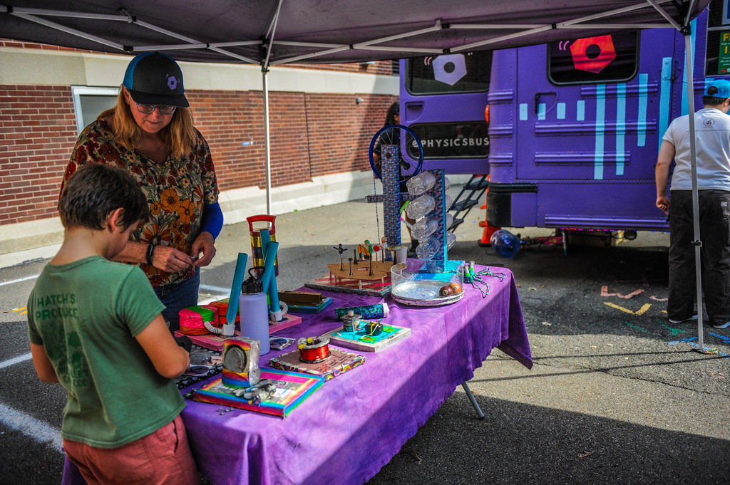 A woman and boy look at items on a table under a tent, with a purple bus parked nearby.