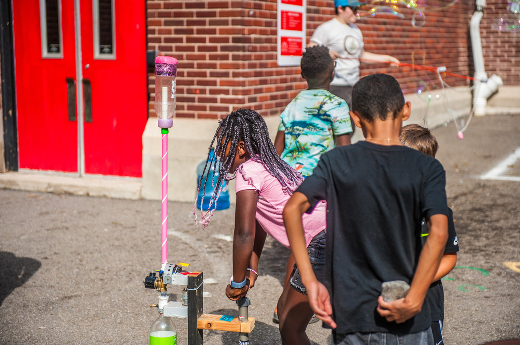 Several children playing with bubbles in front of a red door. A bubble machine is on the left.