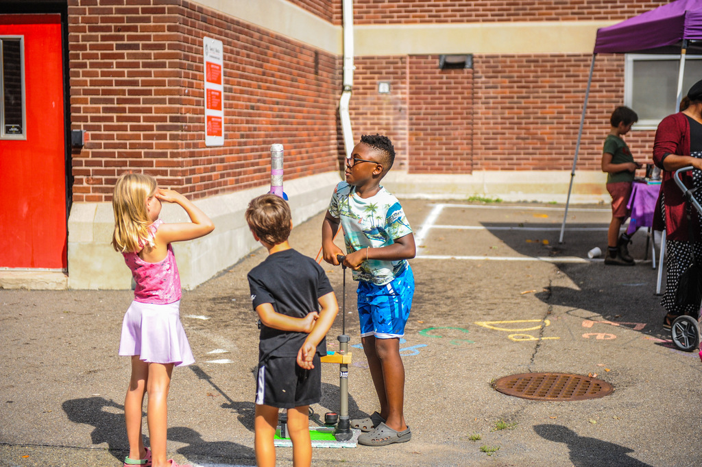Four children play with a toy on a pavement. Brick wall and a tent are in the background.