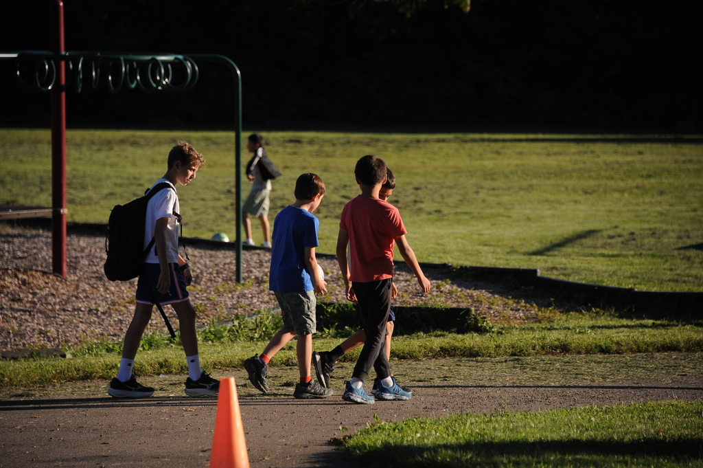 Four boys walk in a park area; one has a backpack. A traffic cone stands on the path.