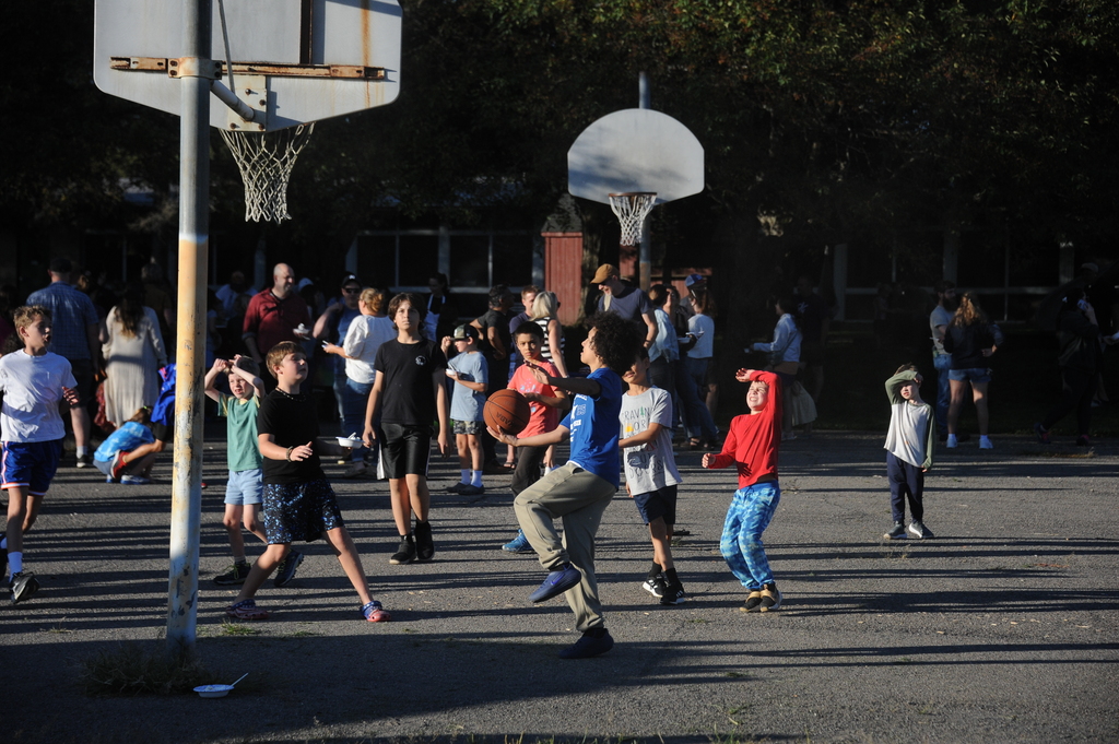 A group of children plays basketball on an outdoor court with trees in the background.
