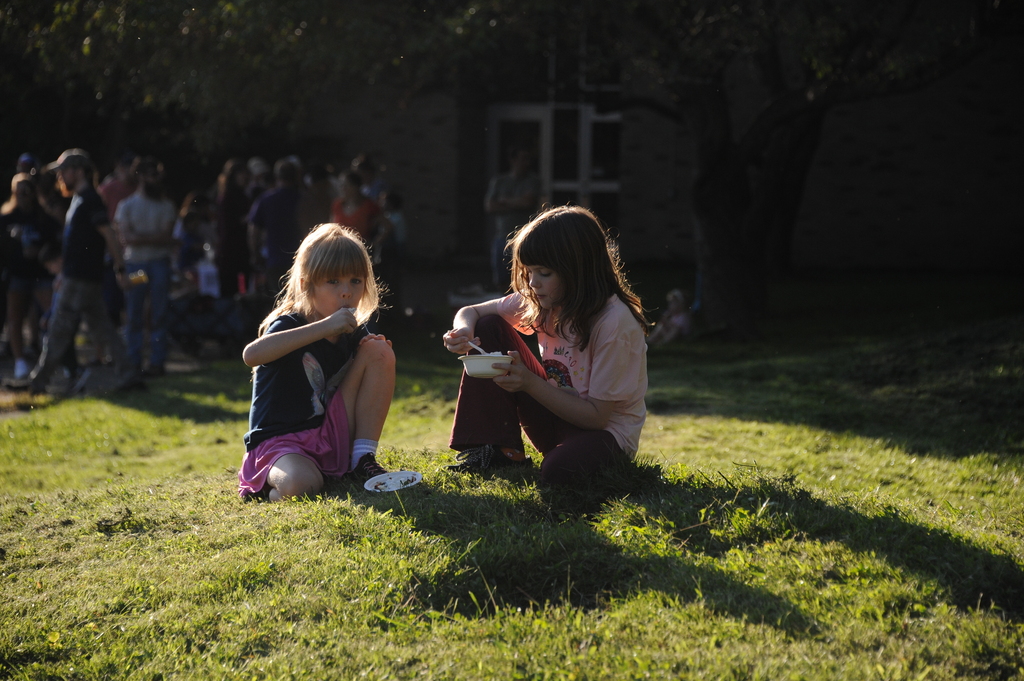 Two girls eating on a lawn with a large crowd of people standing behind them.