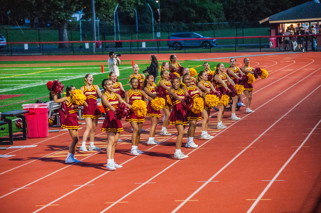 Cheerleaders in maroon and yellow uniforms performing on a red track, with grass and a building in the background.
