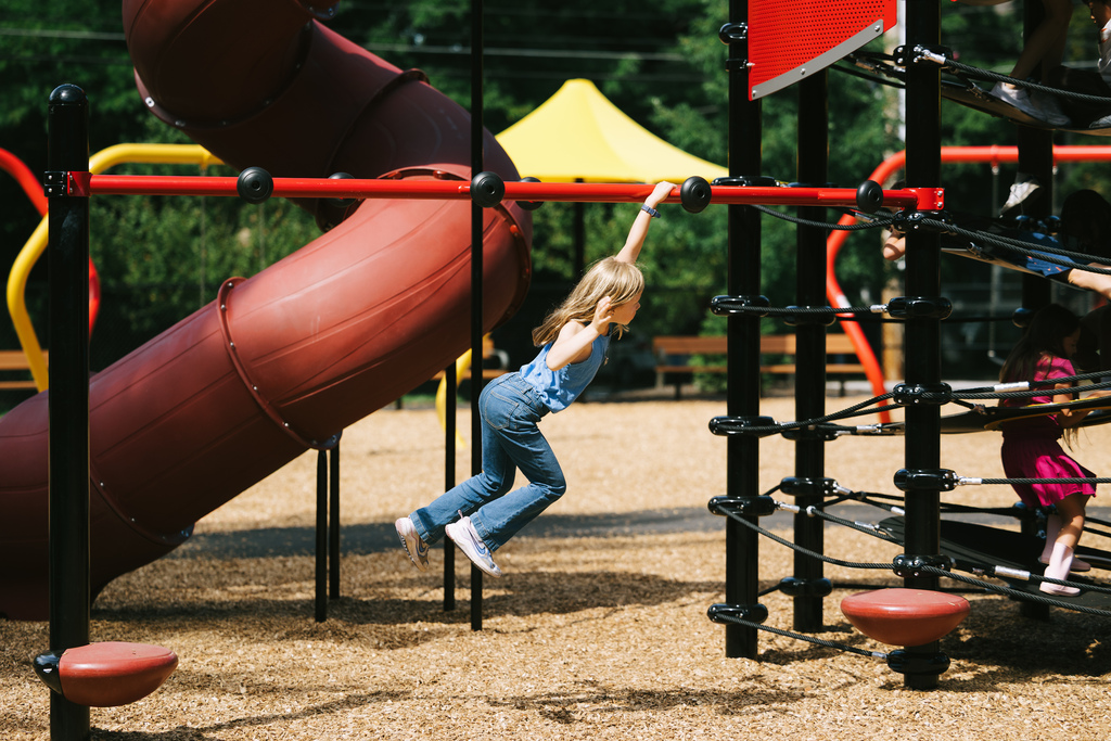 A young child performs a pull-up on a playground climbing structure. A red slide is in the background.
