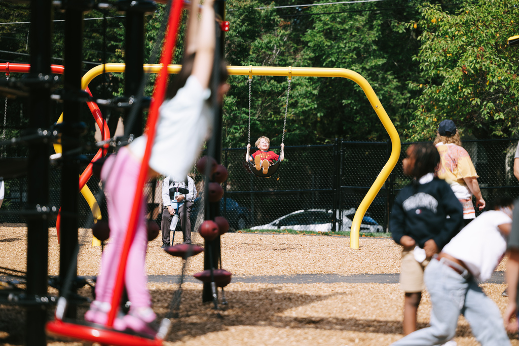 Playground with children. A girl swings and others play near her. Background includes trees and a fence.