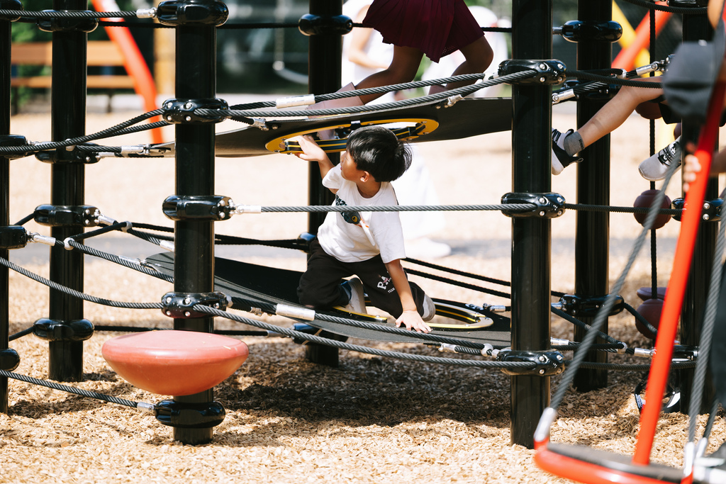 Child on playground equipment, grasping the structure for support, with other kids nearby. Ground covered in gravel.