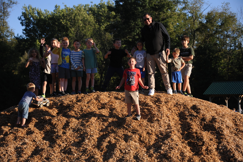 A group of children and an adult stand atop a mound of dirt, with trees in the background.