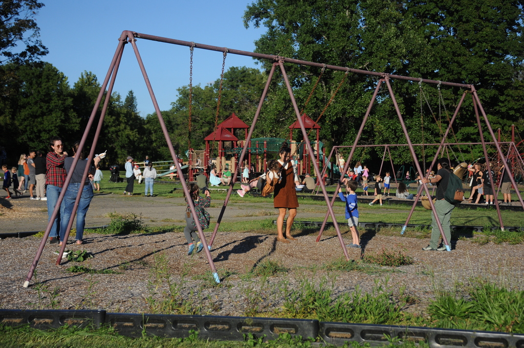 A playground with several swings and a small group of people standing and playing, with trees in the background.