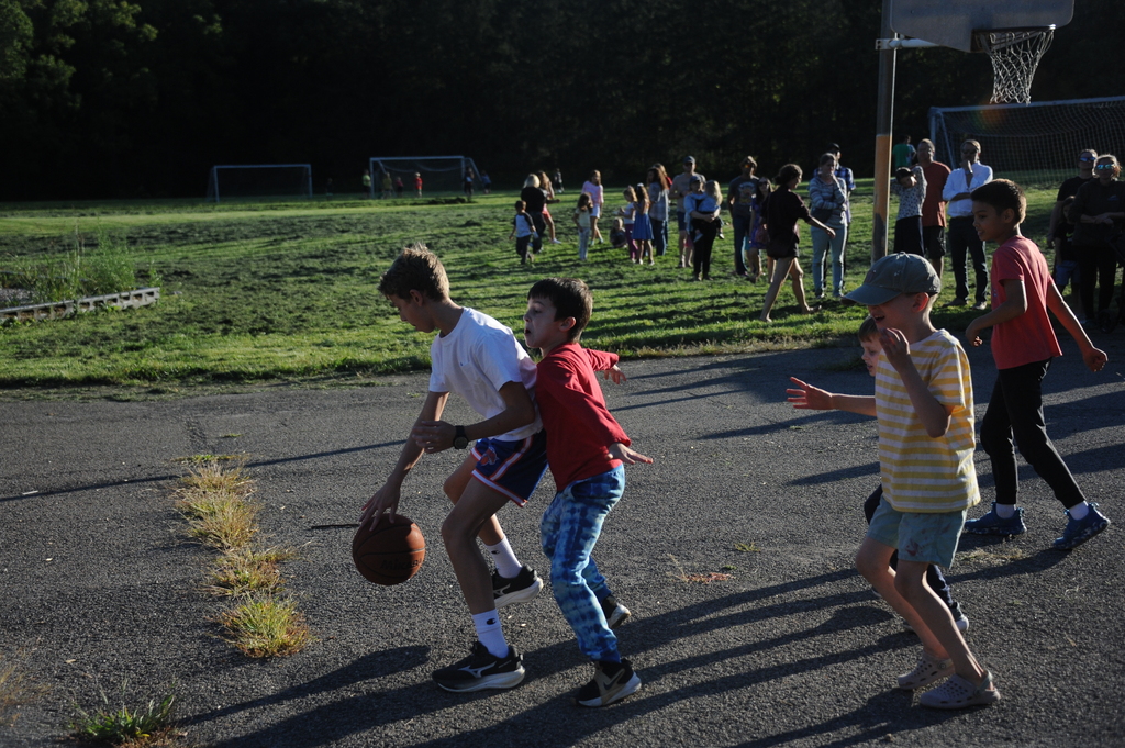 Multiple children playing basketball on a court, others standing in the background on a grassy field.