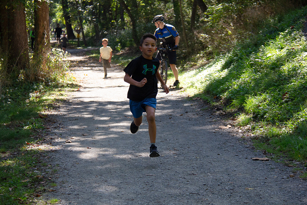 A boy runs on a gravel path with a man in a helmet on a bicycle behind him.