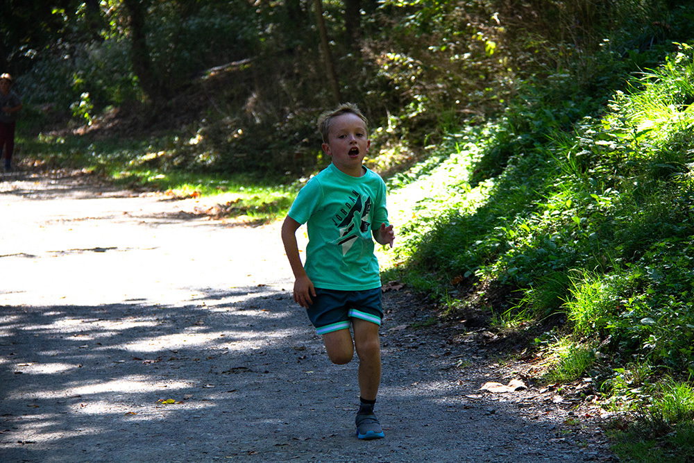 A young child runs on a gravel path in a forest with sunlight and shadows.