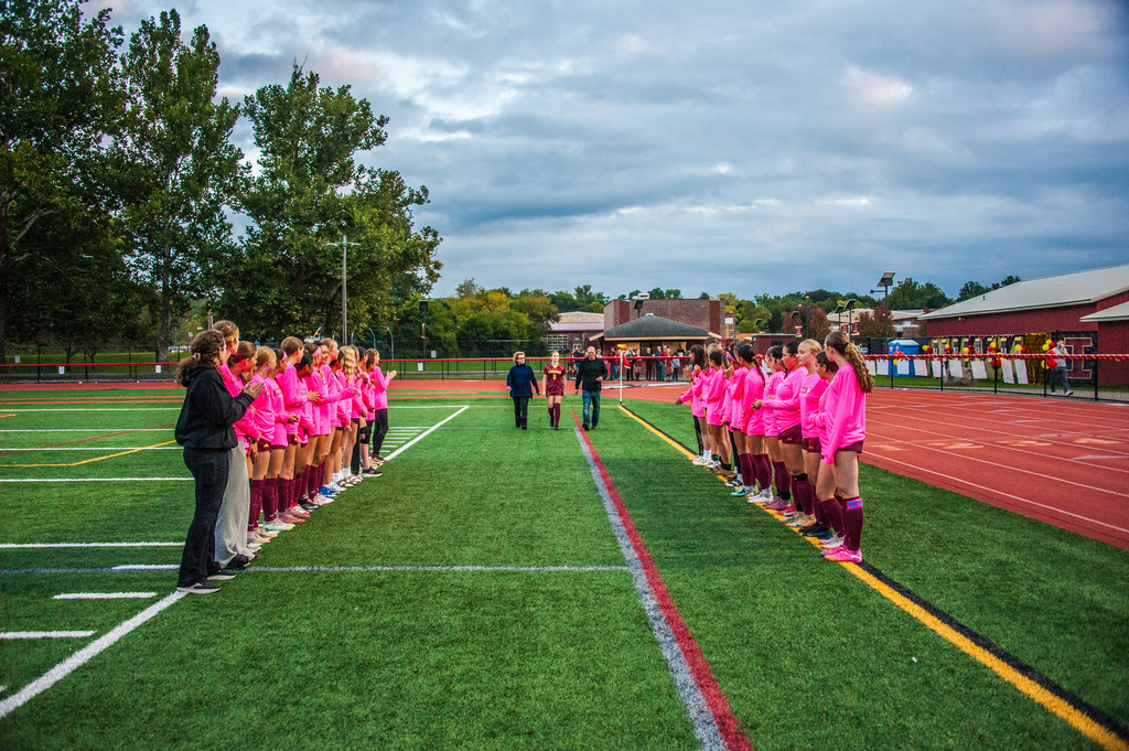 Players in pink shirts and shorts stand on a field; a coach walks between teams; trees and a building in the background.