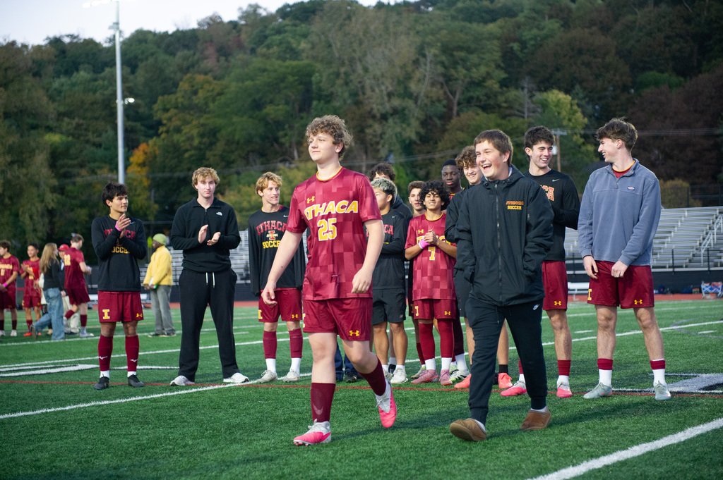 Athletes on a field. One player wears a maroon jersey. Trees and bleachers in the background.