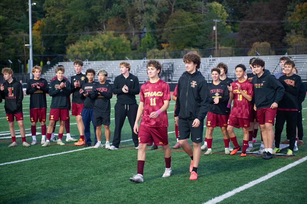 A soccer team stands on a field with green turf, wearing red and black uniforms. Trees and a building are in the background.