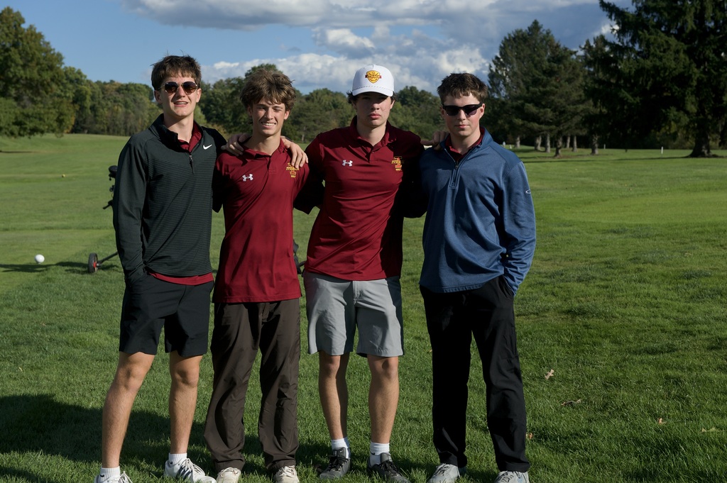 Four men on a golf course. One wears sunglasses and a cap, while others wear shirts in maroon and navy.