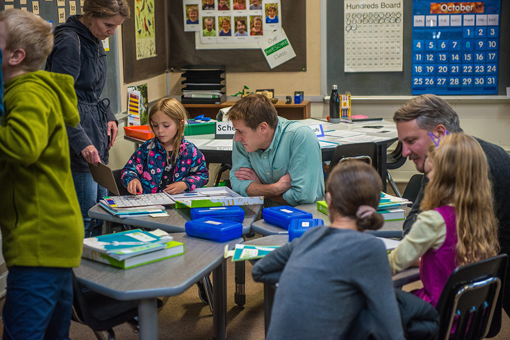 A group of children and adults in a classroom, seated around desks, engaging in activities.