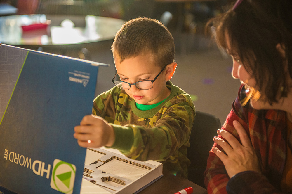 A boy wearing glasses looks at a puzzle box while a woman watches