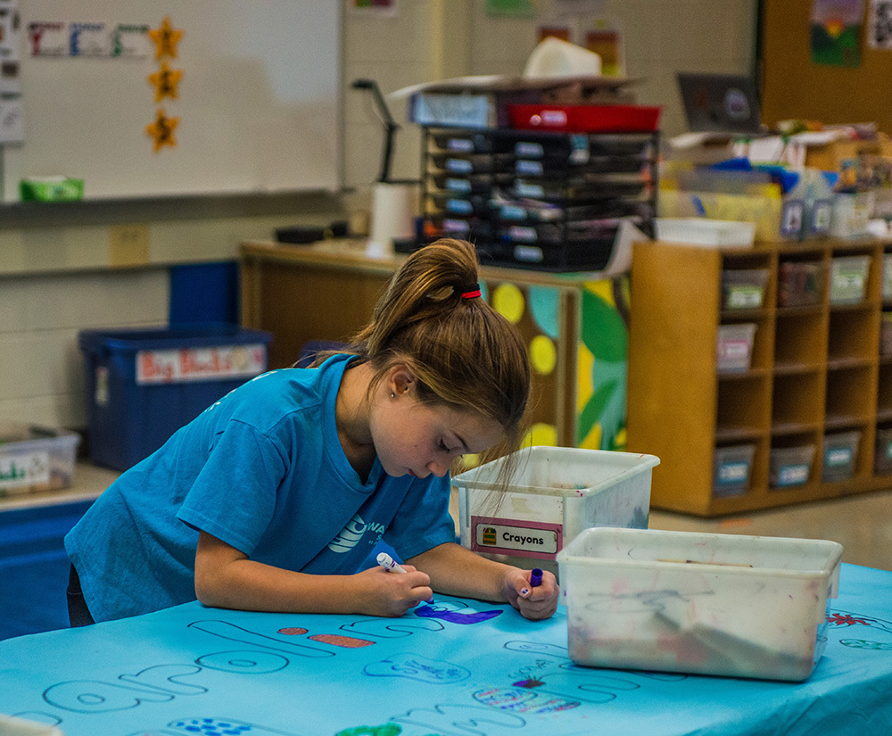 A girl in a blue shirt is painting a table inside a classroom.