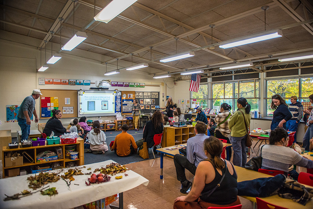Classroom with students and adults sitting and standing listening to a teacher