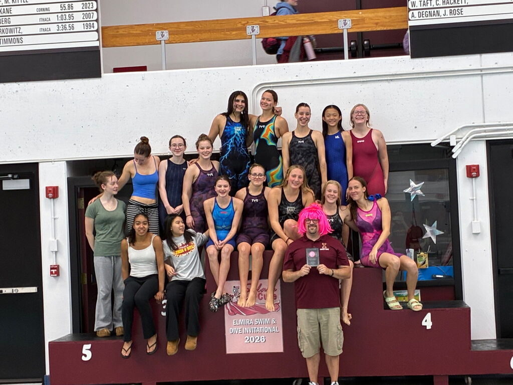 Group of women in swimwear posing on a podium. One woman holds a trophy. A red and white wall with a balcony is behind them.