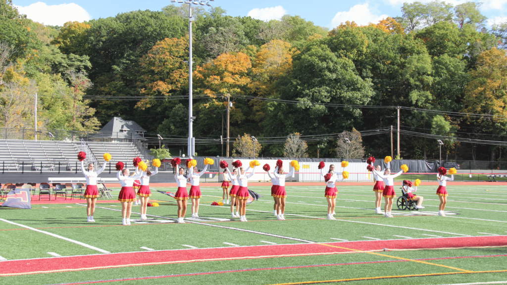 A group of cheerleaders with red and white uniforms performing on a football field with trees in the background.