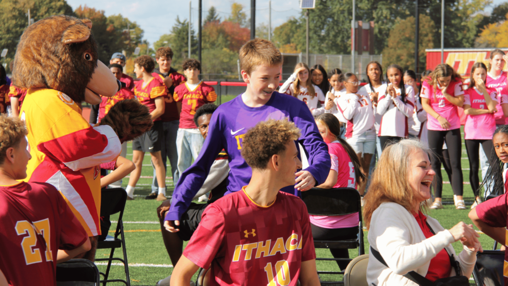 Group on field with mascot. Students in maroon and white. Person in purple with "ITHACA" shirt.