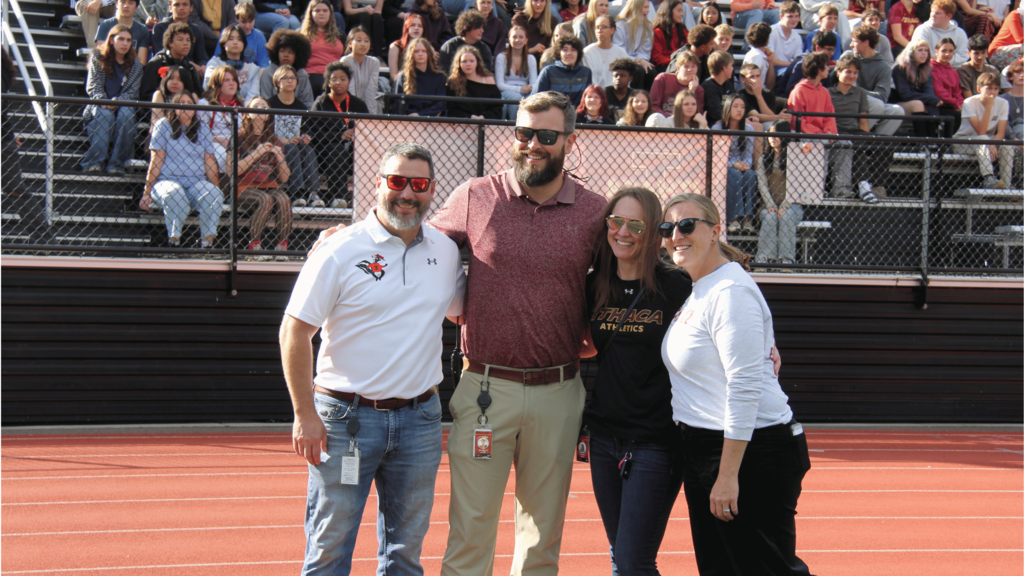 Four people in a stadium, smiling, wearing sunglasses, standing on a track, with a crowd in the background.