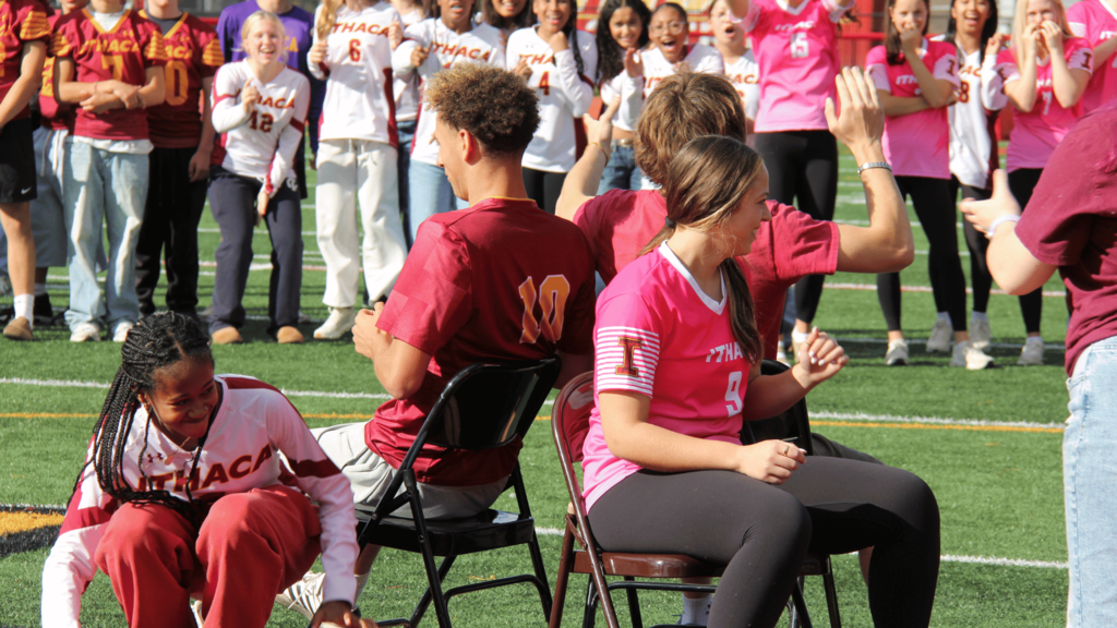 Athletes in maroon and pink jerseys sit in chairs on a field. A crowd in maroon and pink stands behind them, clapping.